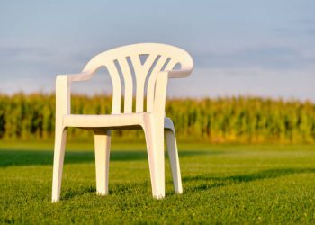 Empy white plastic garden chair standing on a green lawn in front of a cornfield in evening light. Seen in Bavaria, Germany in August.